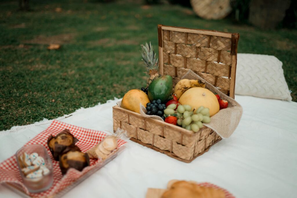 Picnic Accessories - Category Page 4 A delightful picnic scene showcasing a fruit basket and pastries on a white blanket outdoors.